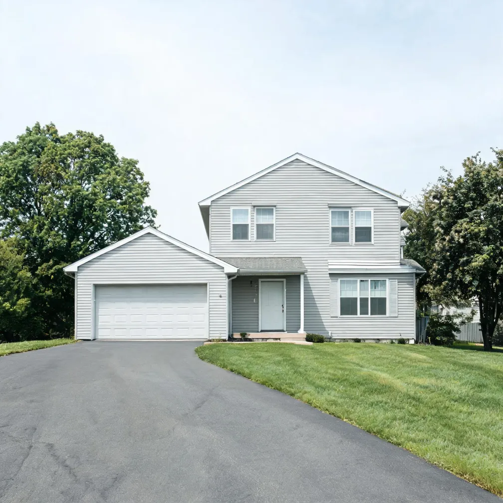 A two-story light gray vinyl-sided house with an attached two-car garage, white front door, and an asphalt driveway curving across a mowed lawn, flanked by leafy green trees under a hazy sky.