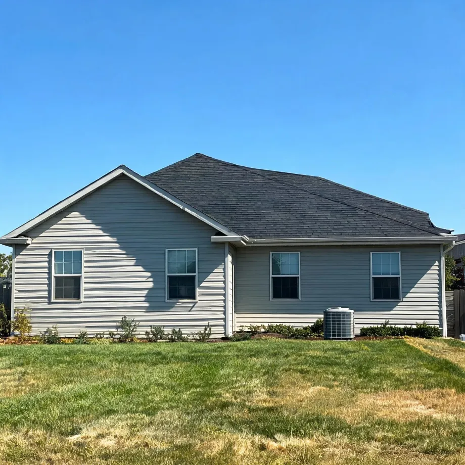 Rear exterior of a single-story house with gray vinyl siding, dark shingled roof, several windows, an air conditioning unit, and a patchy green lawn under a clear blue sky.