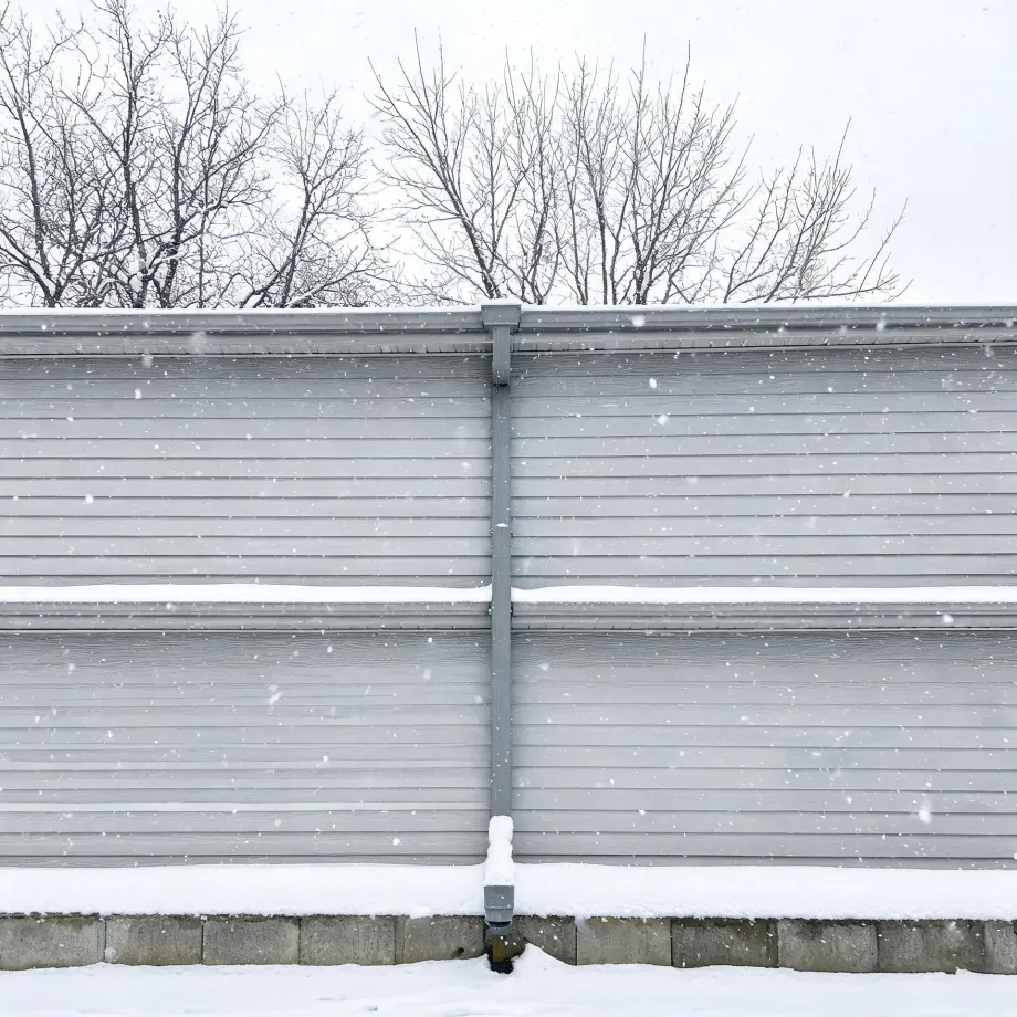 Exterior wall of a building clad in gray horizontal vinyl siding with a vertical downspout at center, a concrete block foundation below, snow on the ground and ledges, and bare trees against a pale sky during falling snow.