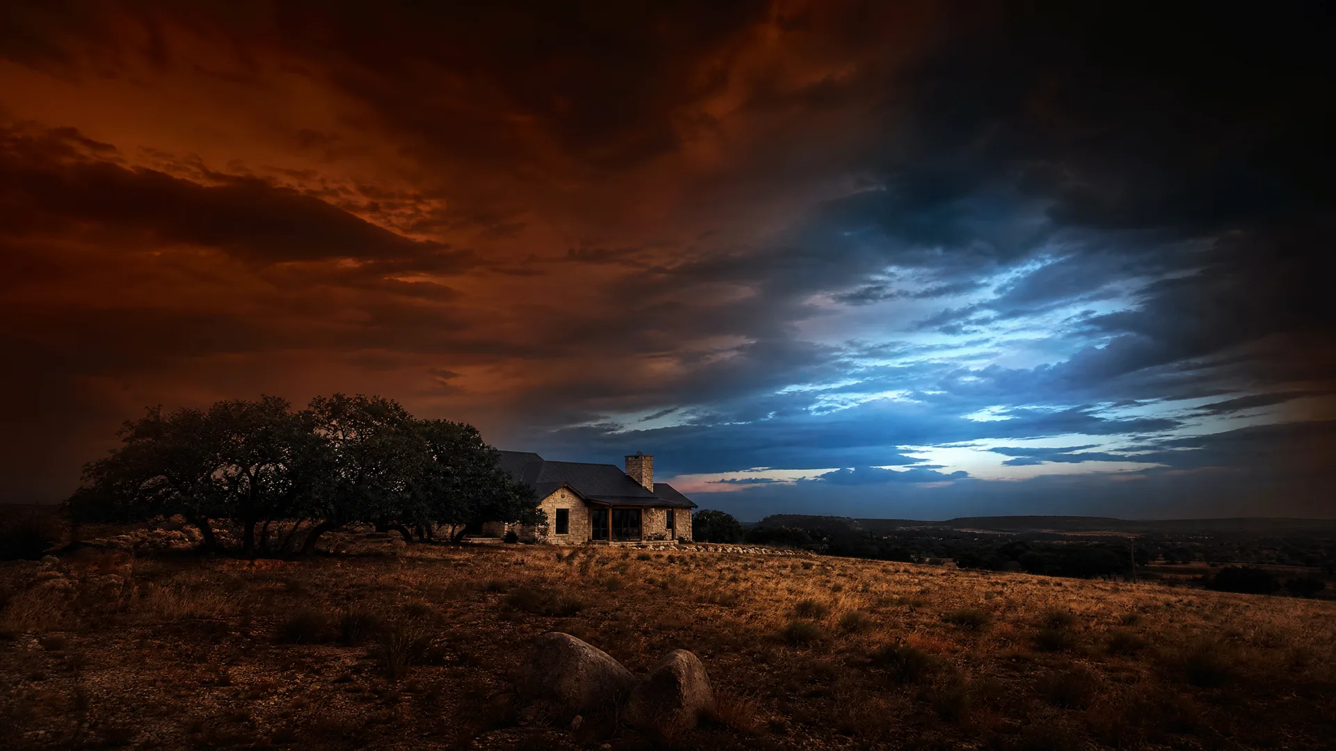 A small stone house with a metal roof and chimney stands beside a cluster of low trees on a dry grassy ridge, under a dramatic dusk sky with orange clouds on the left and blue clouds on the right.