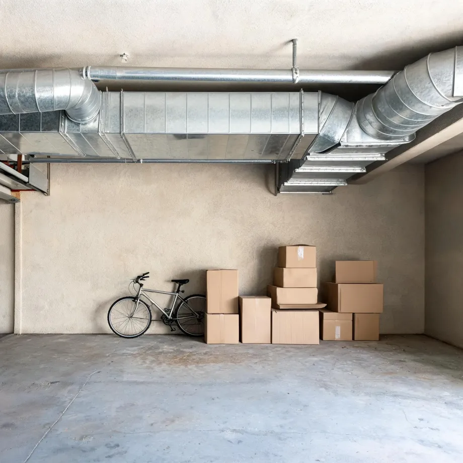 An empty concrete-floored garage or basement room with exposed galvanized rectangular and round ductwork along the ceiling, a bicycle leaning against the beige wall, and a group of stacked cardboard boxes.
