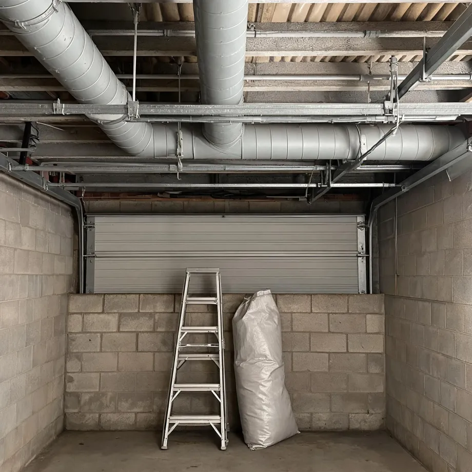 Interior of a concrete block garage with a closed sectional door, exposed ceiling ductwork and conduit, a folding aluminium stepladder and a large white bag on the floor.