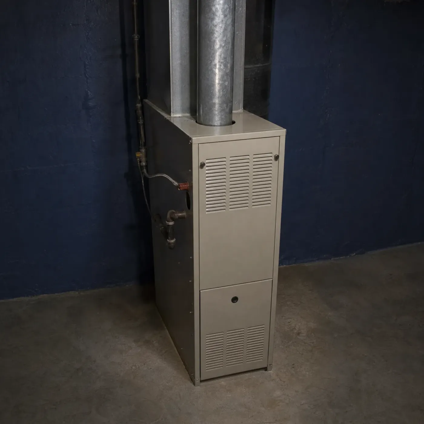 A beige sheet-metal furnace with louvered access panels stands against a dark blue painted basement wall on a concrete floor, with metal flue ducts rising from its top and gas piping along the wall.