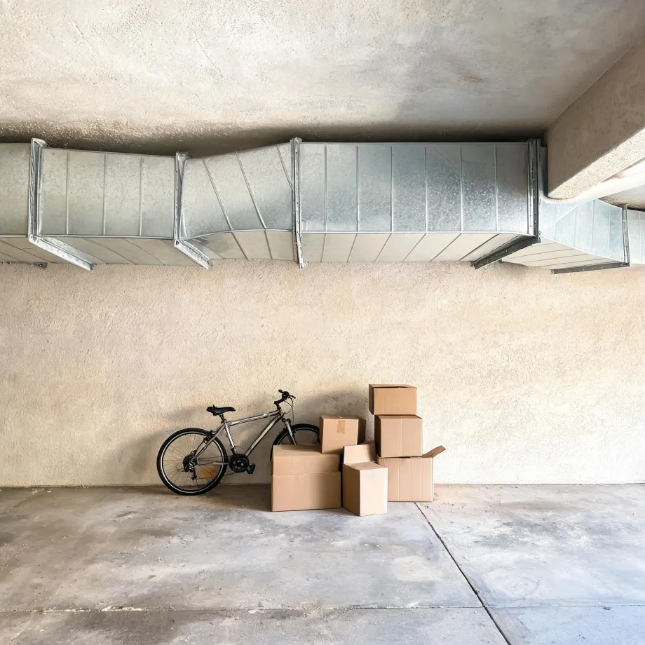 A bicycle leans against a stucco wall in a concrete-floored garage beside a stack of cardboard boxes, with galvanized metal ductwork running along the ceiling above.