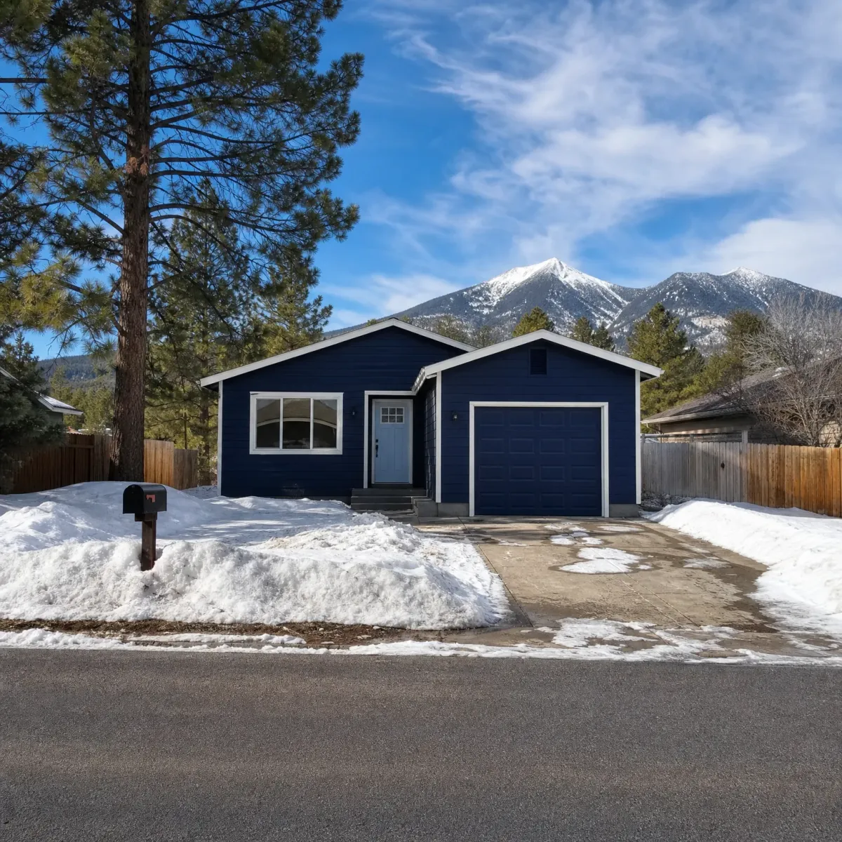 A single-story navy blue house with white trim, a one-car garage and a concrete driveway, surrounded by snow, pine trees, a wood fence, and snow-capped mountains in the background.