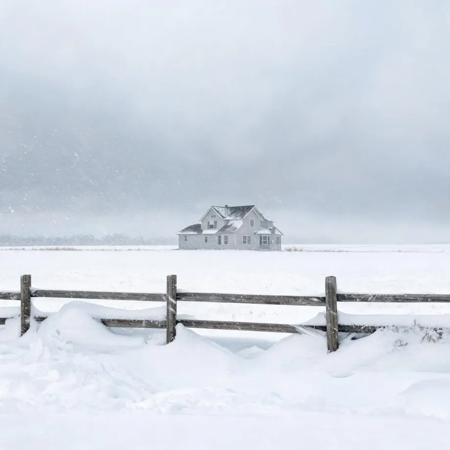 A two-story light-colored house stands alone in a snow-covered field during falling snow, with a weathered wooden post-and-rail fence and drifts in the foreground under a pale gray sky.