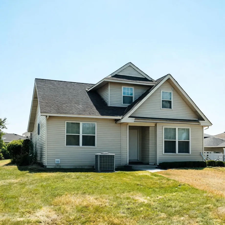 A two-story house with beige horizontal vinyl siding, gray shingled gables and dormer, white-framed windows, a central entry door, and an outdoor air conditioning unit, set on a partly dry lawn under a clear blue sky.