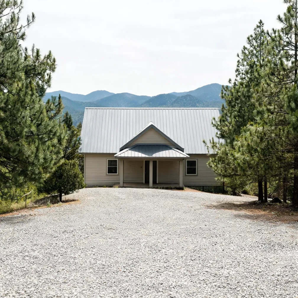 A single-story beige house with a metal gable roof and covered front entry, at the end of a wide gravel driveway, flanked by pine trees with forested mountain ridges in the background.