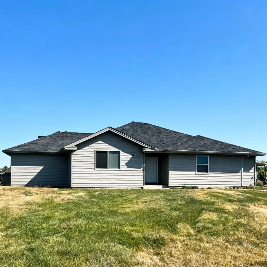 Single-story house with gray lap siding and a dark shingled roof, with a grass lawn in front under a clear blue sky.
