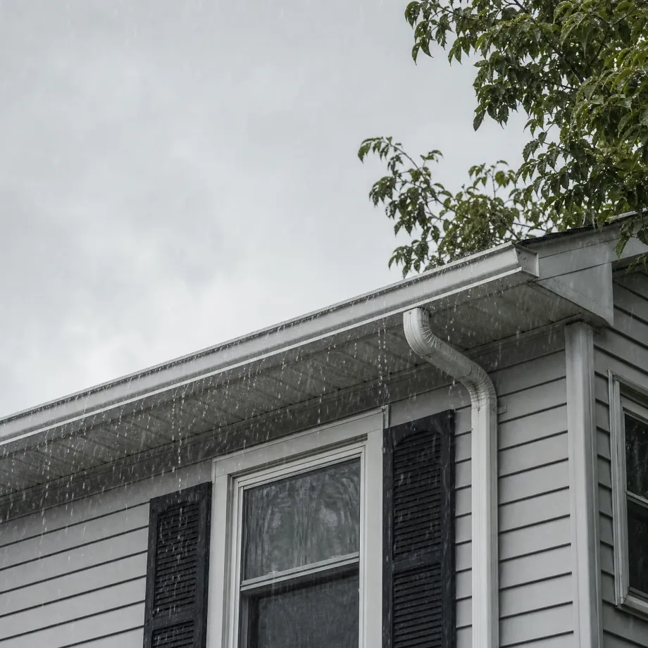 Rain falls past the roof edge and white gutter with a downspout on a gray horizontal-sided house with black shutters, under an overcast sky with tree branches at upper right.