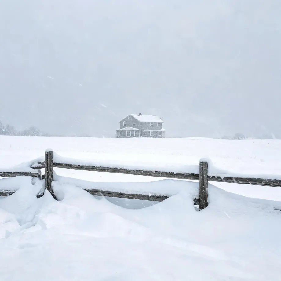 A two-story house stands on a snow-covered field during falling snow, with a partly buried wooden post-and-rail fence in the foreground and a pale overcast sky.