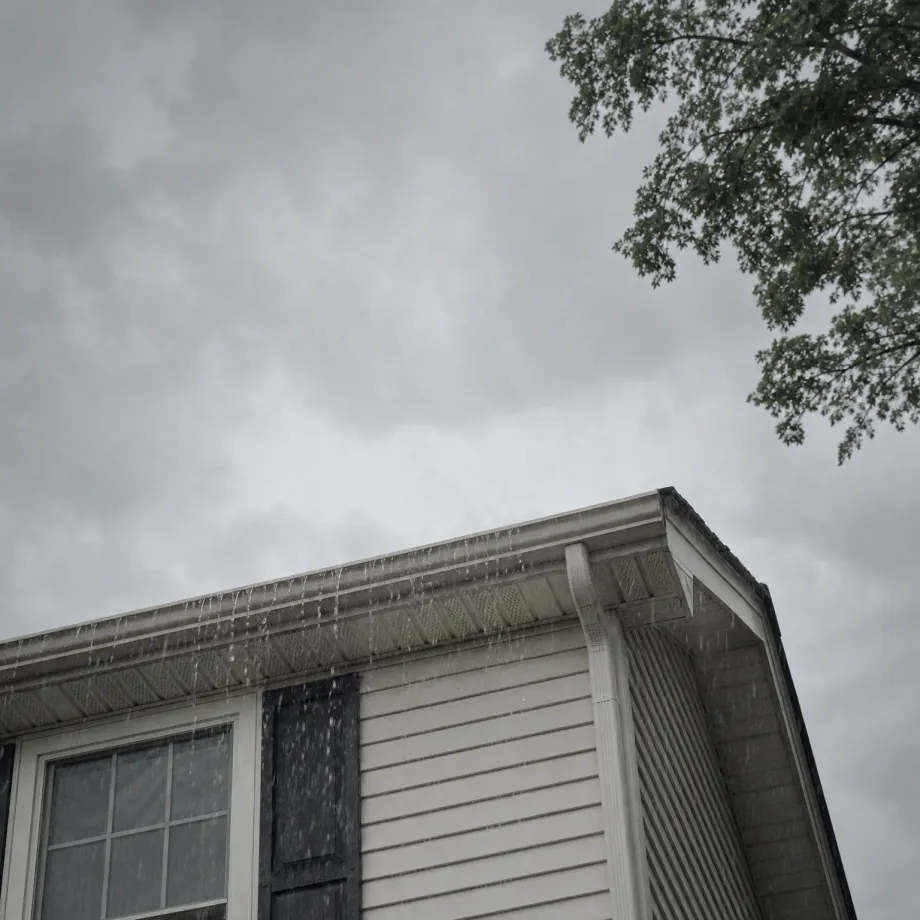 Upward view of the corner of a white horizontal-sided house with water streaming off the roof edge and gutter under an overcast gray sky, with tree branches at the upper right.