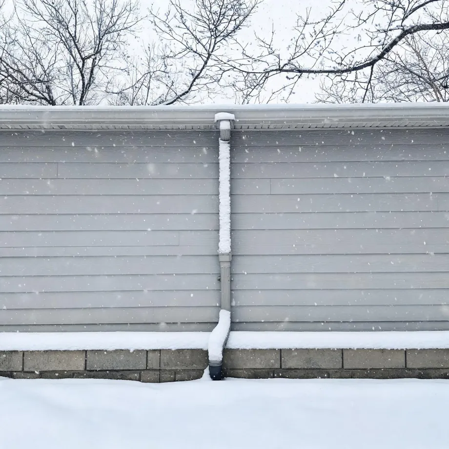 Exterior wall of a building with light gray horizontal lap siding and a concrete block foundation, with a snow-covered downspout running down the center and falling snow, bare trees above the roofline.