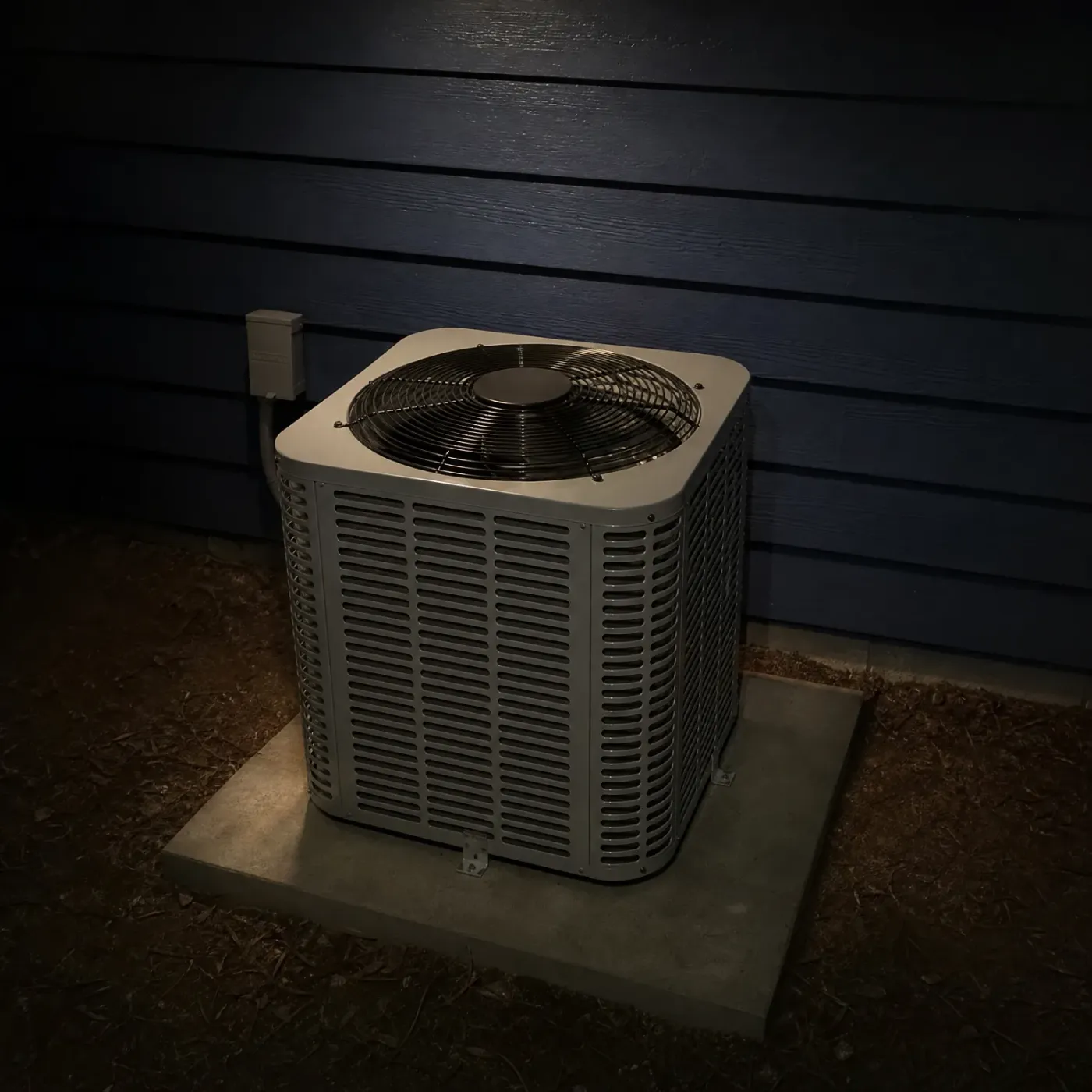 An outdoor air conditioning condenser unit on a concrete pad beside a dark blue horizontal lap-sided wall at night, with a small electrical disconnect box and conduit on the wall above it and mulch surrounding the pad.