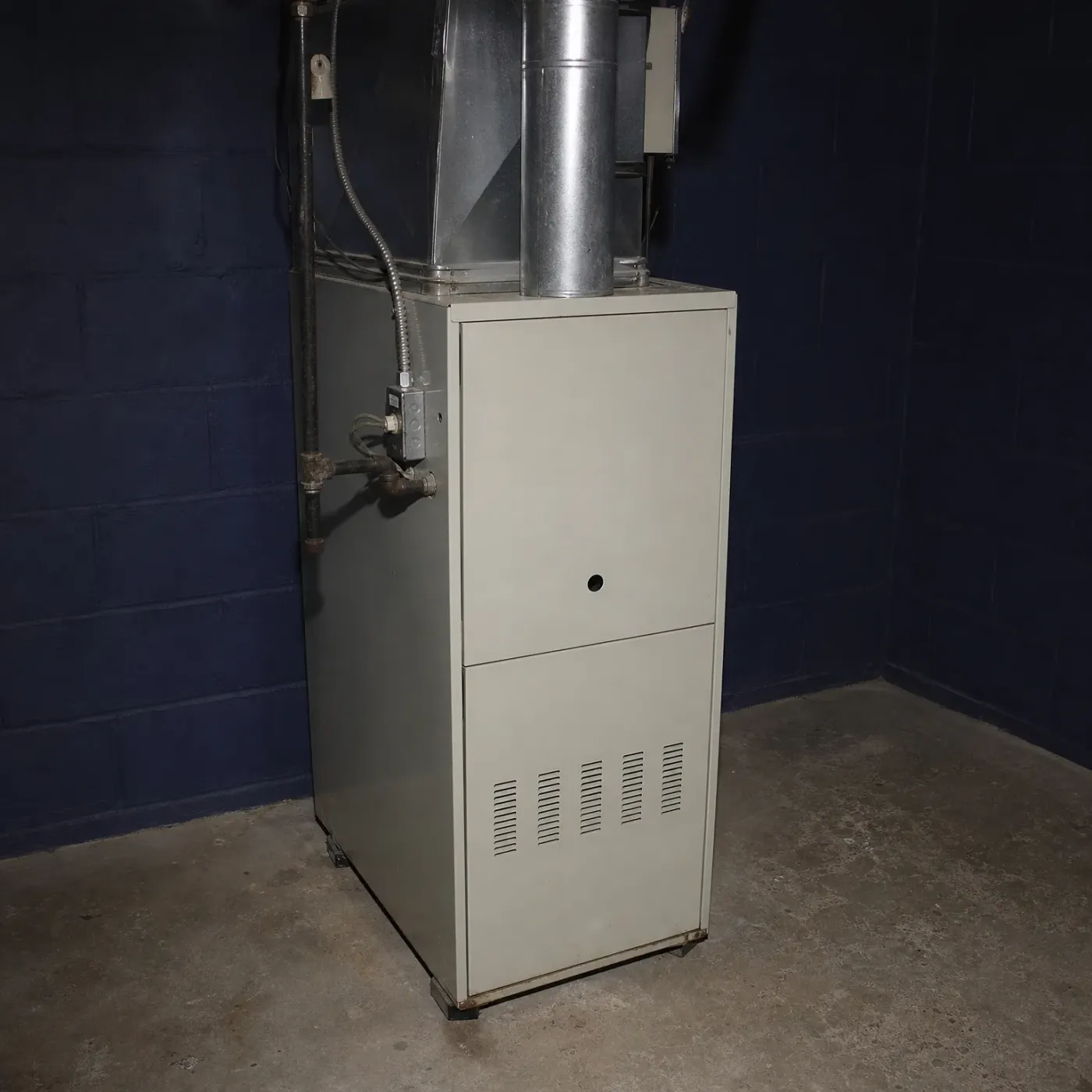 A beige sheet-metal furnace with a louvered lower panel stands on a concrete floor against a dark blue painted concrete block wall, with metal ductwork, a flue pipe, gas piping and an electrical box above and beside it.