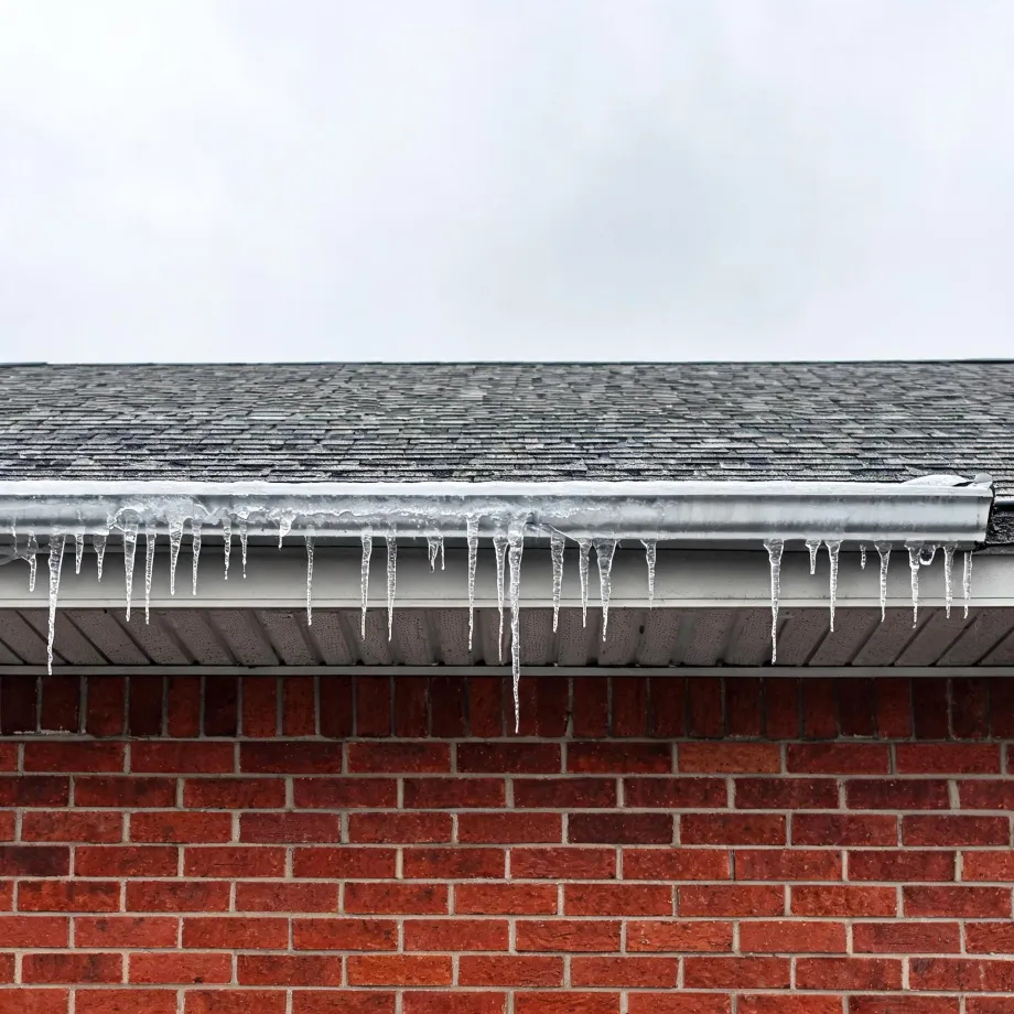 Icicles hanging from a white gutter along the edge of an asphalt-shingle roof above a red brick wall, under an overcast sky.