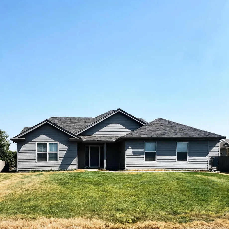 Single-story gray horizontal-lap-sided house with dark shingled gable and hip roofs, white-trimmed windows and a front door, fronted by a partly dry green lawn under a clear blue sky.