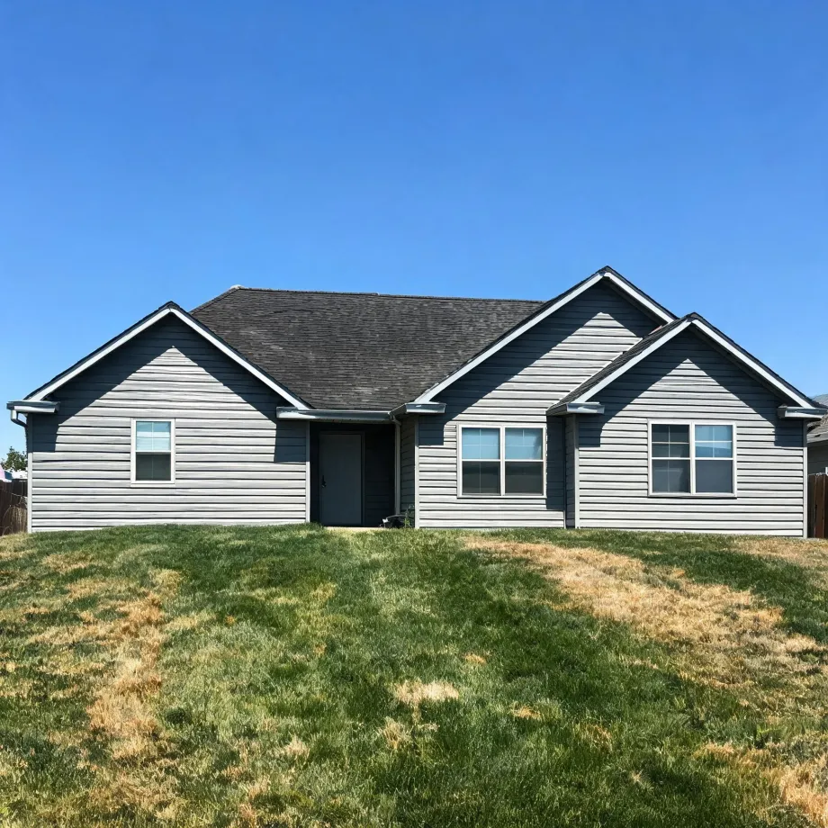 Single-story gray horizontal-siding house with multiple front gables, dark shingle roof, a dark front door and two windows, fronted by a patchy green and brown lawn under a clear blue sky.