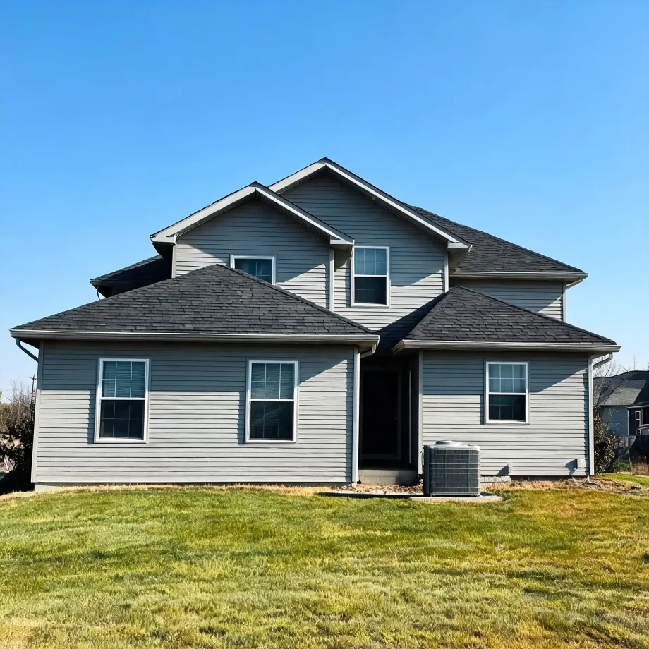Rear view of a two-story gray vinyl-sided house with dark shingled gables, white-trimmed windows, an open doorway, an outdoor air conditioning unit, and a grassy lawn under a clear blue sky.