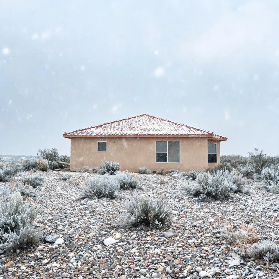A single-story tan stucco house with a red tile roof stands amid snow-dusted desert scrub and a rocky gravel foreground during falling snow.