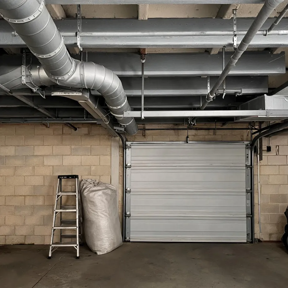 Interior of a garage with concrete block walls, a closed white sectional overhead door, exposed steel beams and metal ductwork on the ceiling, a folding step ladder and a large white bag on the floor.
