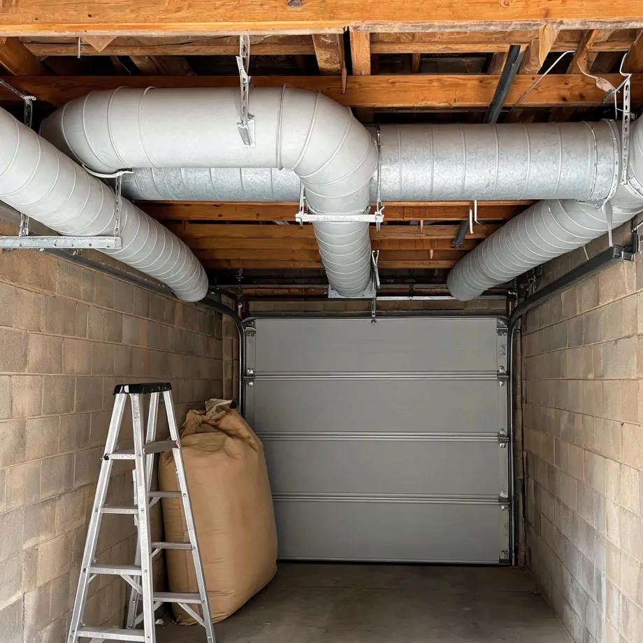 Interior of a concrete block garage with a closed sectional door, large round metal ducts running along the exposed wood joist ceiling, an aluminum step ladder and a tall fabric sack on the floor.