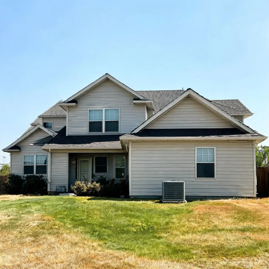 Two-story house with beige horizontal siding, gray shingled gable roofs and a covered front entry, with an outdoor air conditioning unit beside it and a partly dry grass lawn in front under a clear blue sky.