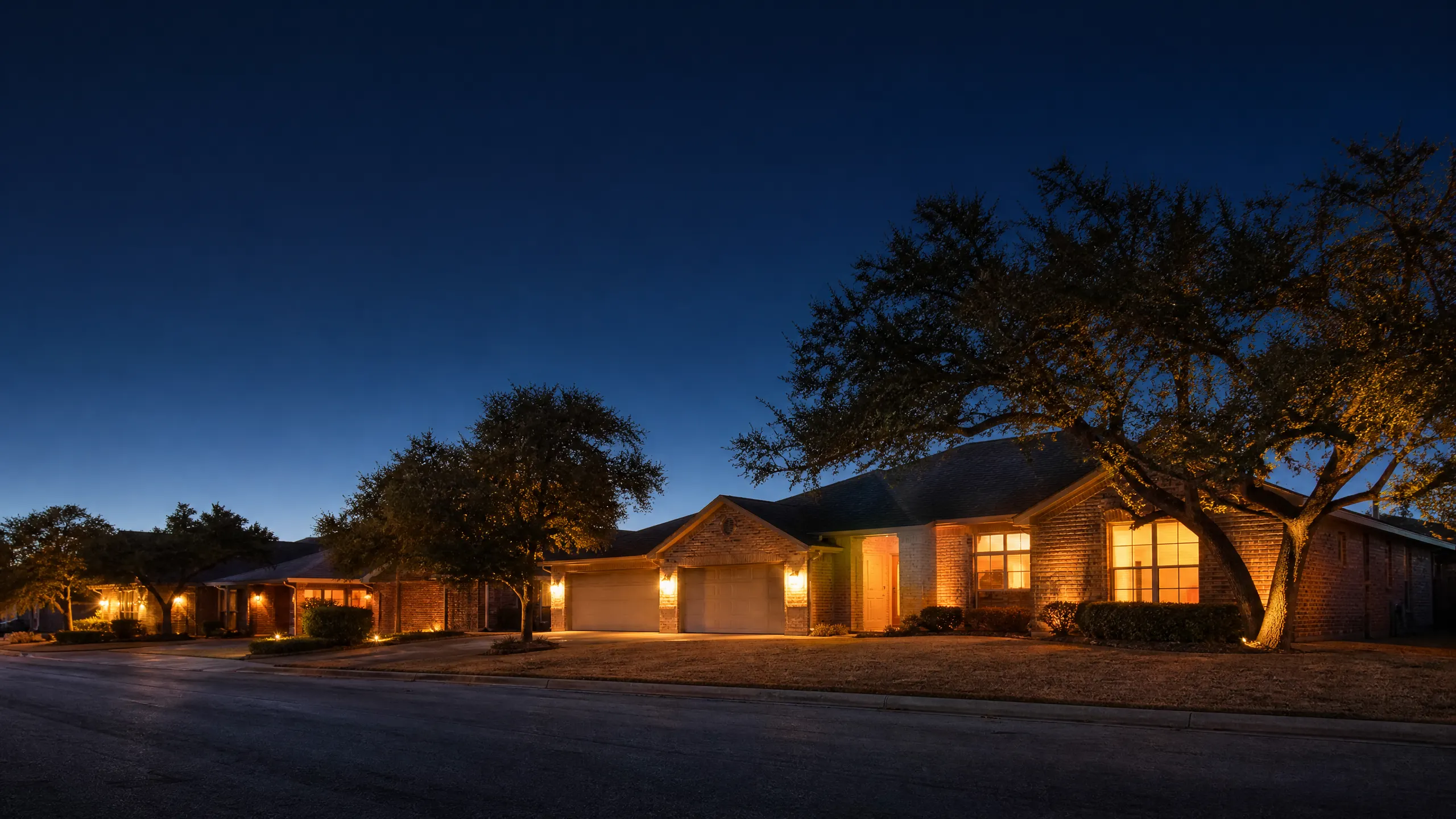 Single-story brick ranch houses with lit garages and large oak trees along a residential street at dusk.