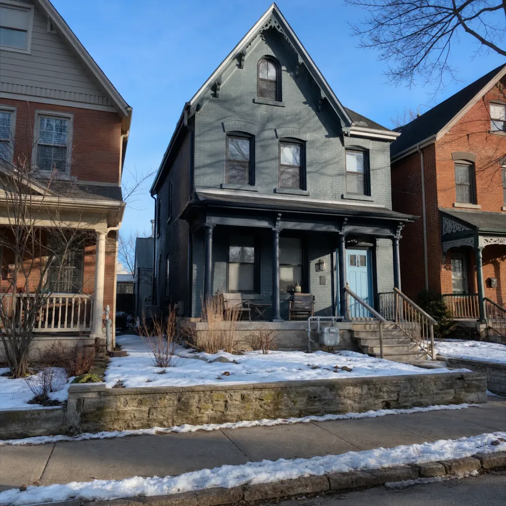 A two-and-a-half-storey gray-painted brick house with a gabled roof, covered porch with columns, and light blue front door, flanked by red brick houses, with snow on the front yard and sidewalk.