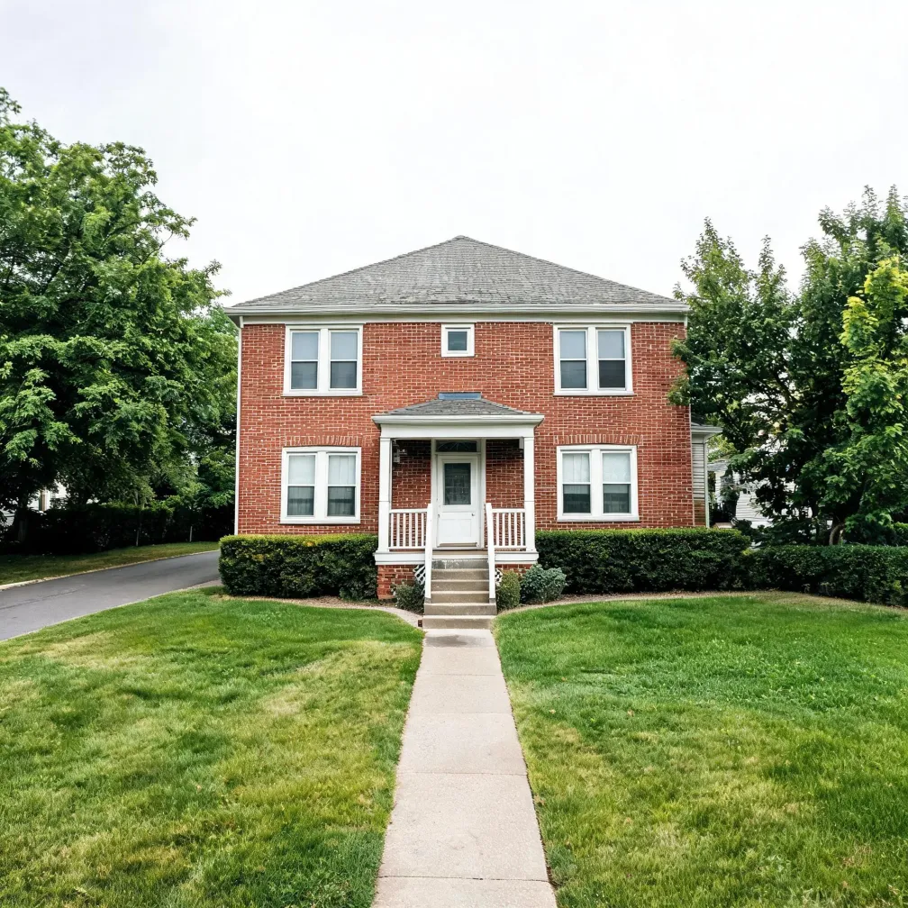 A two-story red brick house with a hipped gray roof, white-trimmed windows, and a small covered front porch with steps leading to a concrete walkway across a green lawn, flanked by trimmed hedges and leafy trees.