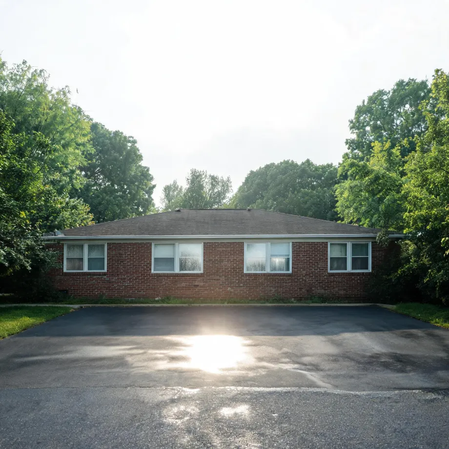 A single-story red brick house with a low shingled roof and several white-framed windows, fronted by a large empty asphalt lot with sun glare, surrounded by green deciduous trees under a bright overcast sky.