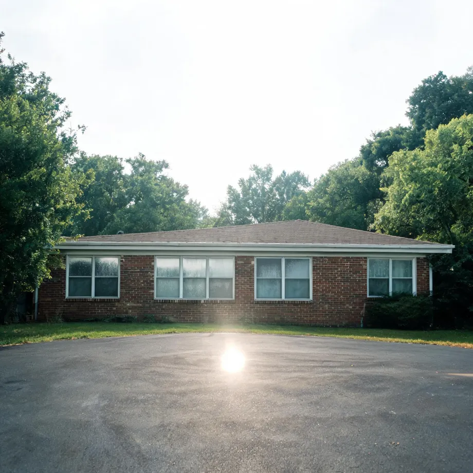 A single-story red brick building with a hipped shingle roof and several white-framed windows, fronted by a large empty asphalt lot with sun flare, surrounded by green trees under a bright hazy sky.