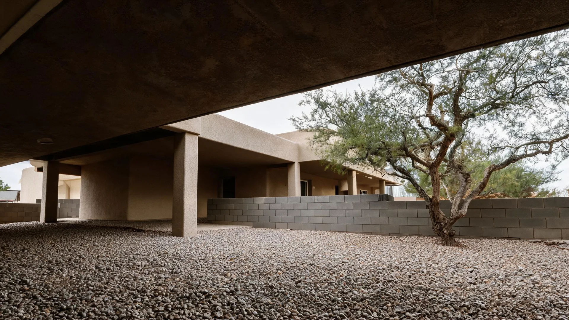 View from beneath a concrete overhang across a gravel yard toward a single-story stucco building with square columns, a gray concrete block wall, and a large bare-branched tree at right.