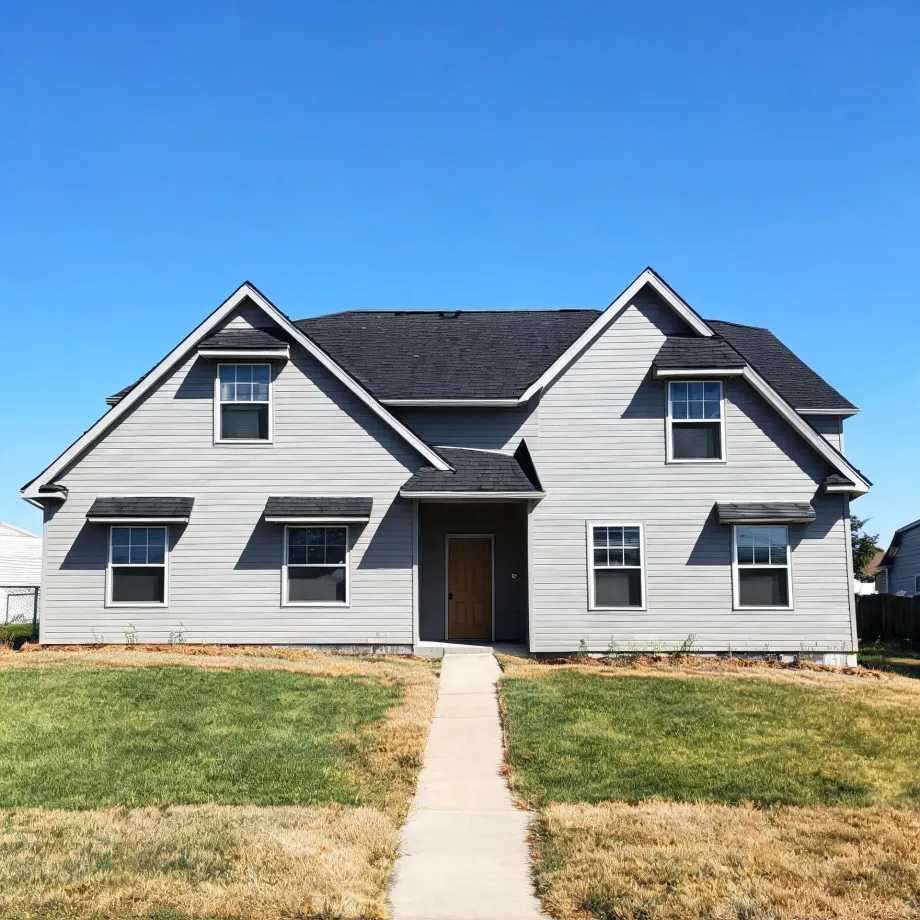 A two-story gray horizontal-siding house with dark shingled gable roofs, a brown front door, and a concrete walkway crossing a patchy green and dry lawn under a clear blue sky.