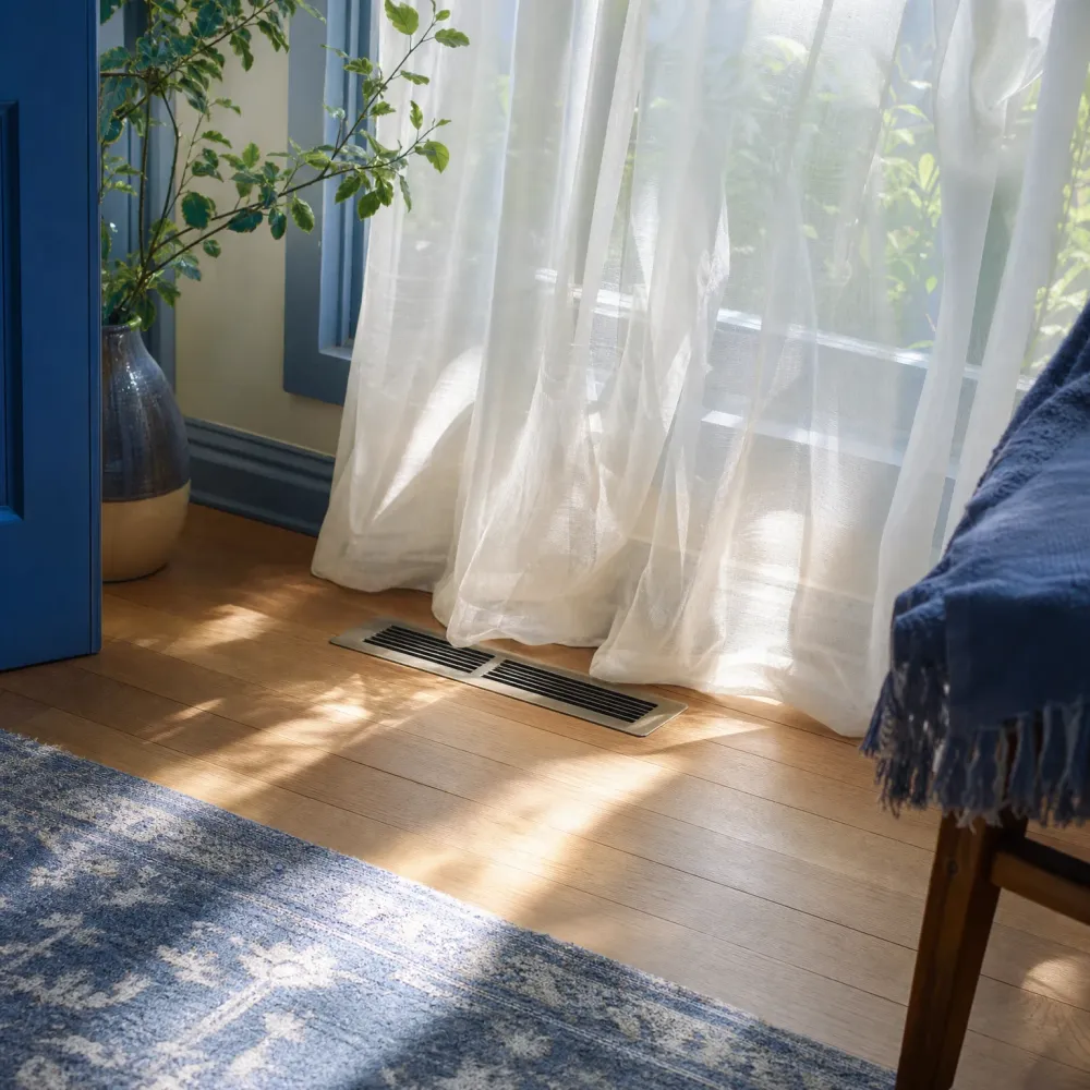 Sunlit wood floor with a metal floor vent near sheer white curtains at a window, a blue-trimmed doorway and tall potted plant at left, a blue patterned rug in the foreground, and a wooden chair draped with a blue fringed throw at right.