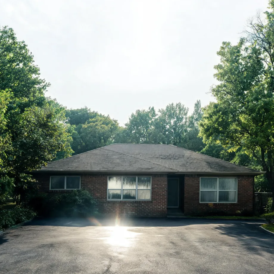 A single-story brick house with a hipped shingle roof, large front windows and a central door, fronted by a wide paved driveway with sun flare, surrounded by green deciduous trees.