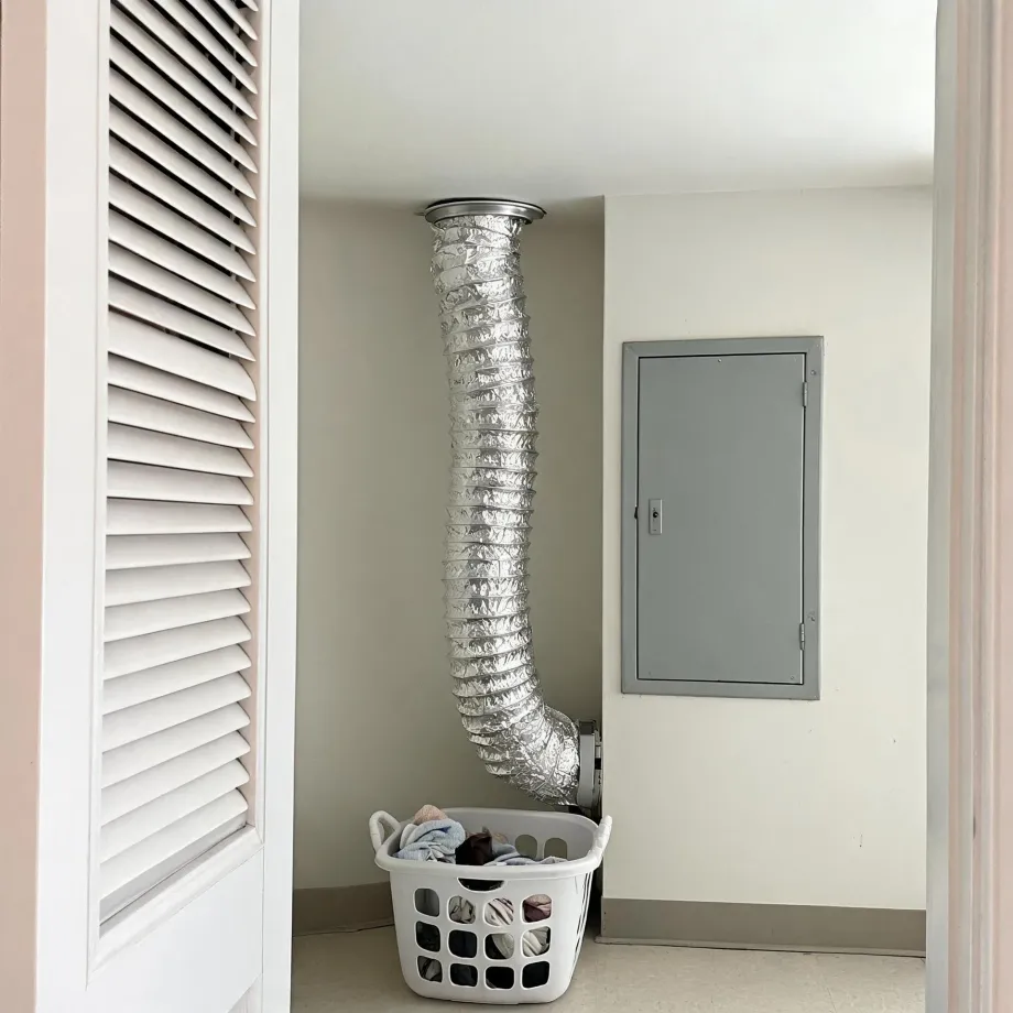 Interior corner of a laundry area where a flexible foil dryer duct runs from a ceiling vent down to a wall connection, with a white plastic laundry basket of clothes below, a gray metal access panel on the wall, and a louvered white door at left.