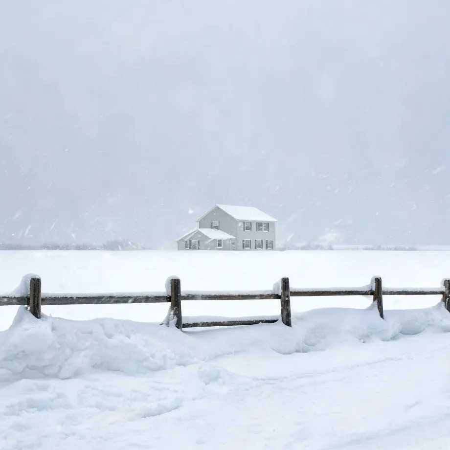 A two-story light-colored house stands alone in a snow-covered field during falling snow, with a snow-topped wooden rail fence crossing the foreground.