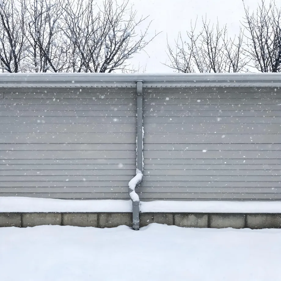 Gray horizontal siding on a low building with a downspout running to a concrete foundation, snow on the ledge and ground, bare trees behind the roof, and snow falling.
