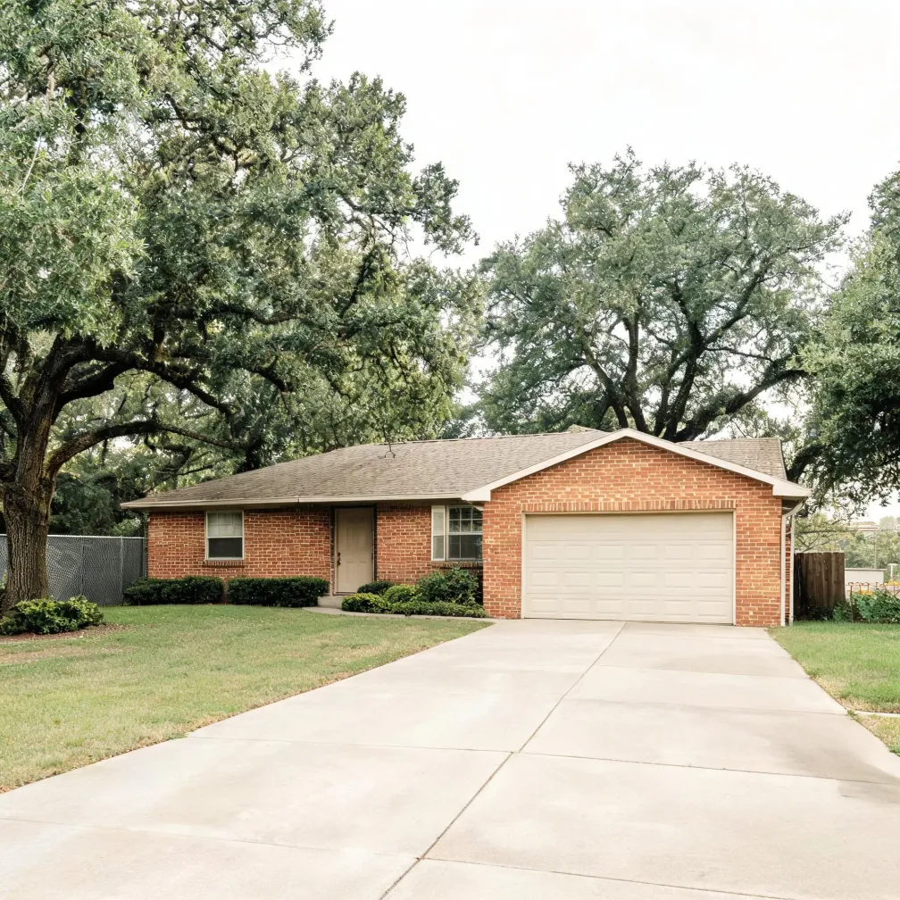 Single-story red brick house with a shingled roof, attached two-car garage with a white door, wide concrete driveway, trimmed shrubs, lawn, and large oak trees overhead.