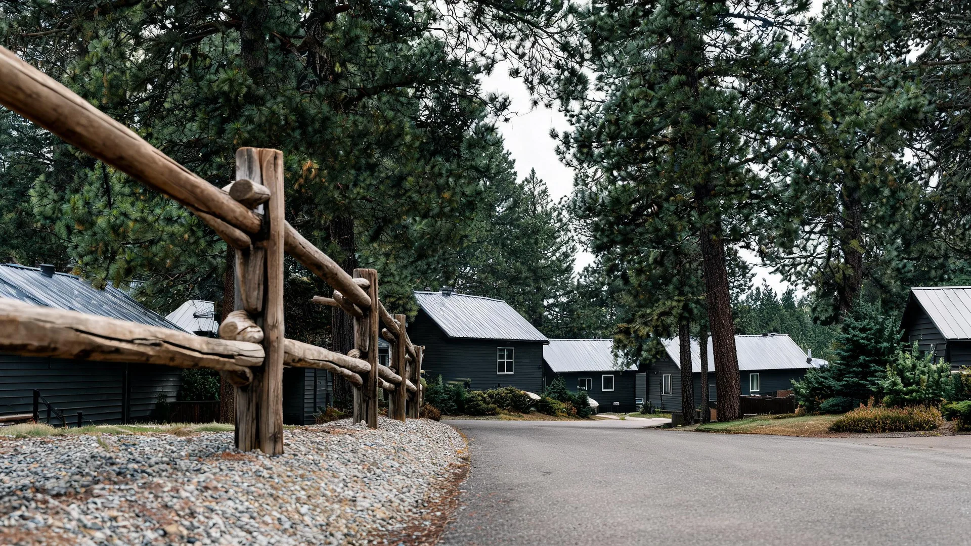 A rustic log rail fence beside a gravel verge and paved road, with dark-sided cabins with metal roofs among tall pine trees under an overcast sky.