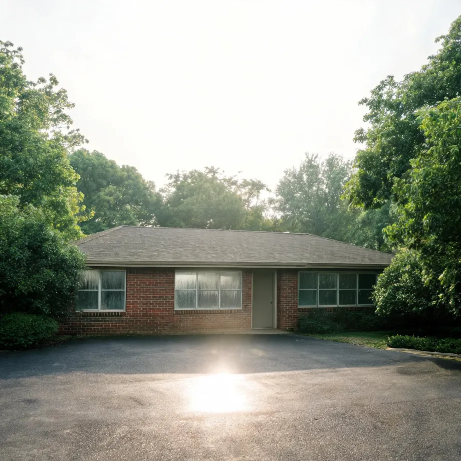 A single-story red brick house with a shingled hip roof, large windows and a central door, fronted by a wide asphalt parking area with sun glare, surrounded by green trees and shrubs.