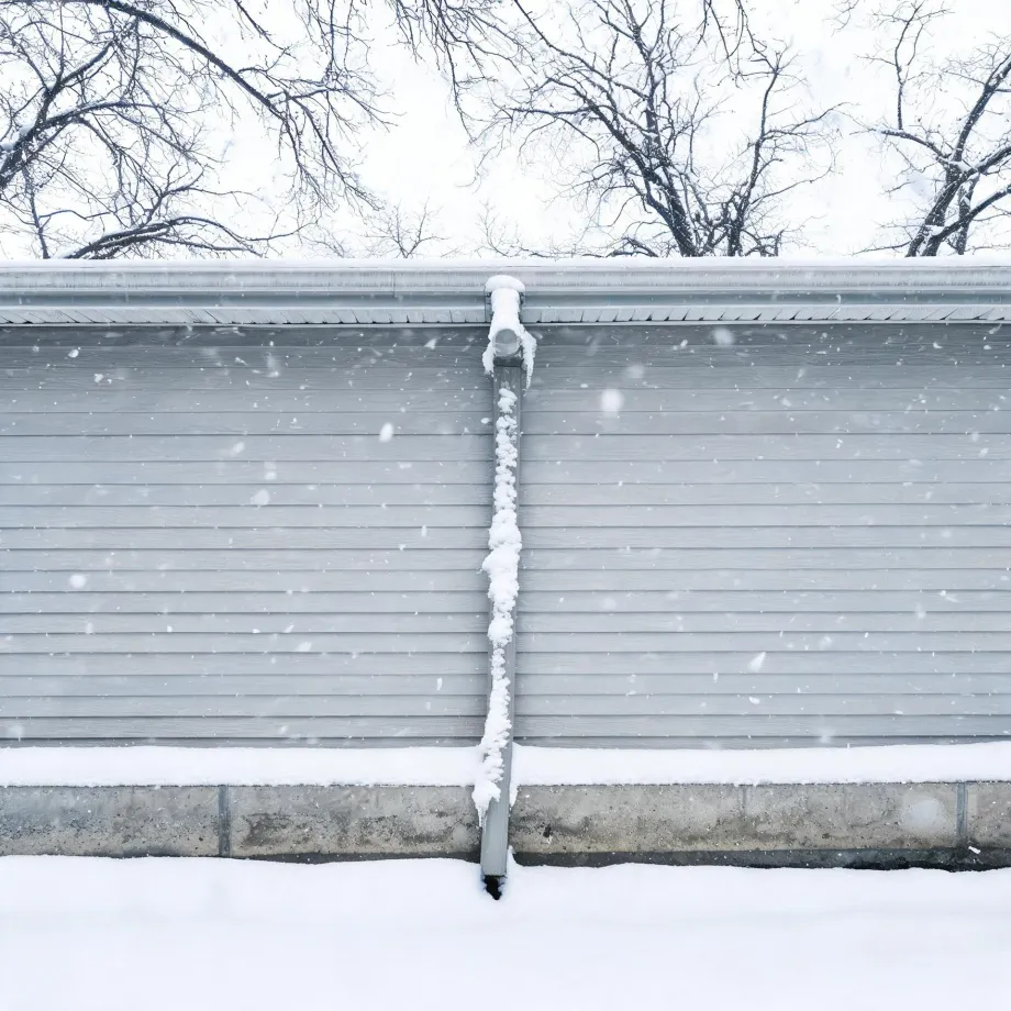 Exterior wall of a building with gray horizontal siding and a concrete foundation, a snow-covered downspout running down the center, falling snow, snow on the ground and roof edge, and bare trees against a white sky.
