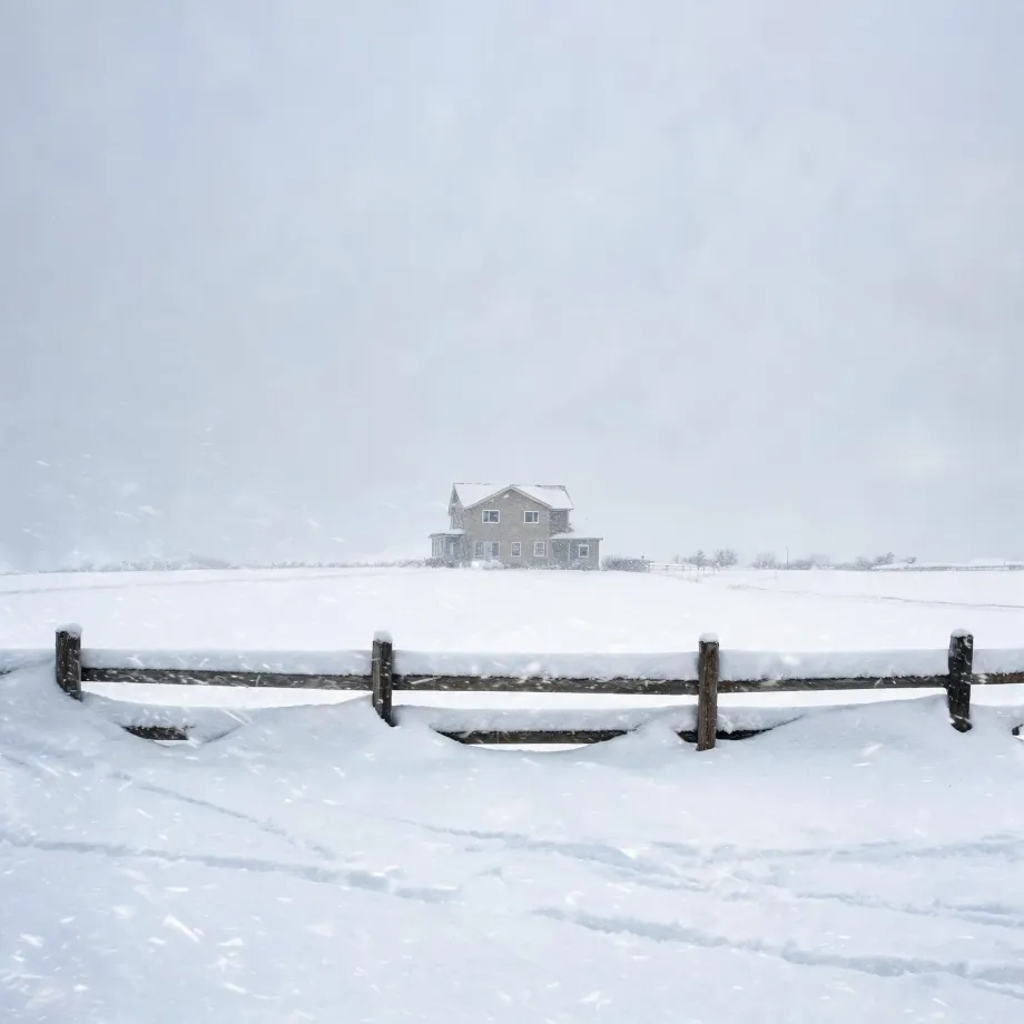 A two-story house stands on a flat snow-covered field in falling snow, with a partly buried wooden post-and-rail fence crossing the foreground under a pale gray sky.