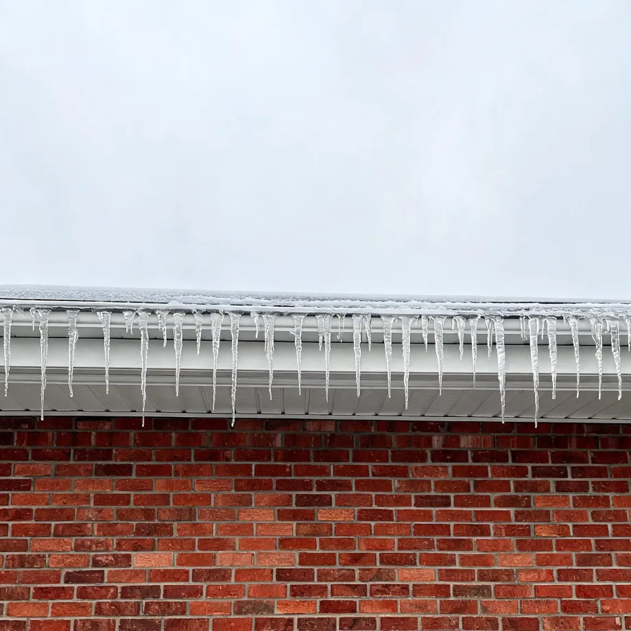 A row of icicles hangs from a white gutter and eave above a red brick wall, with snow on the roof edge and an overcast pale sky above.