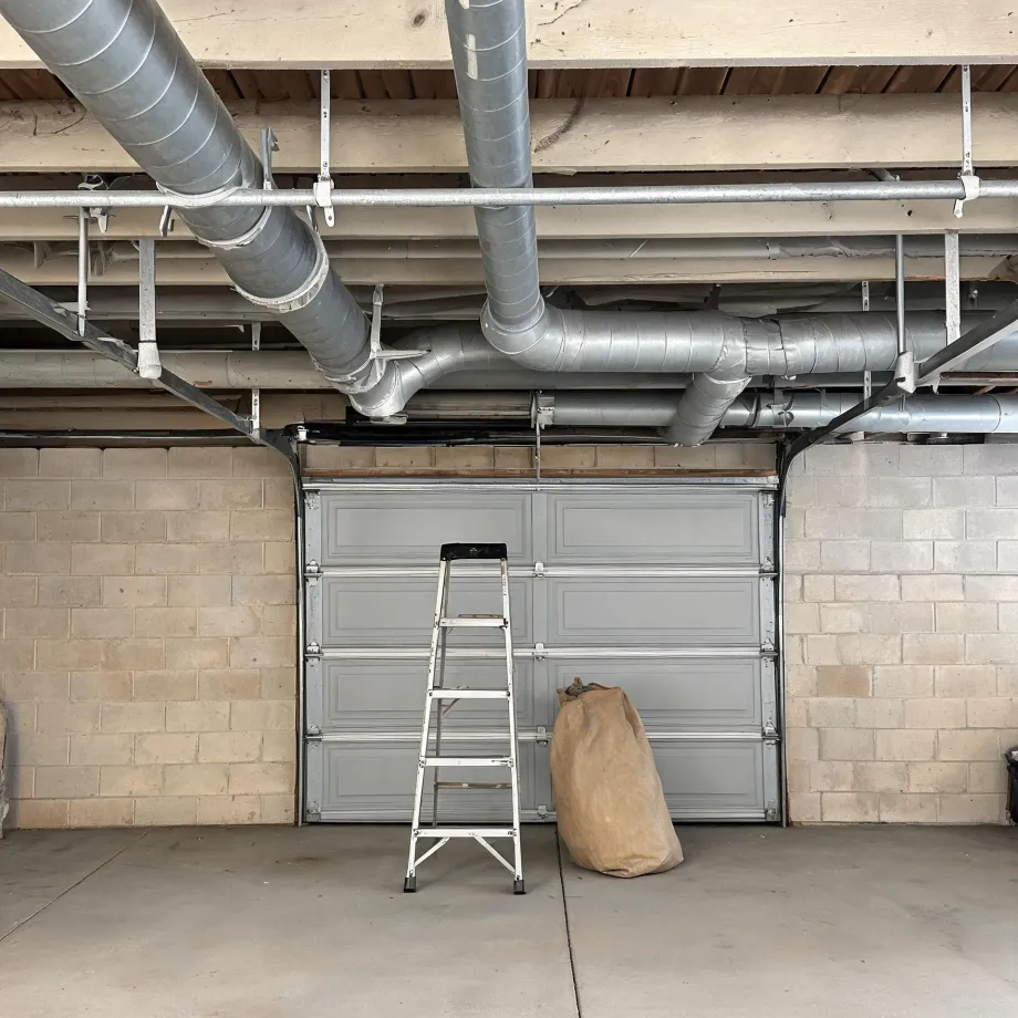 Interior of a concrete block garage with exposed galvanized ductwork and pipes under a wood joist ceiling, a closed sectional garage door, a folding step ladder, and a large brown sack on the concrete floor.