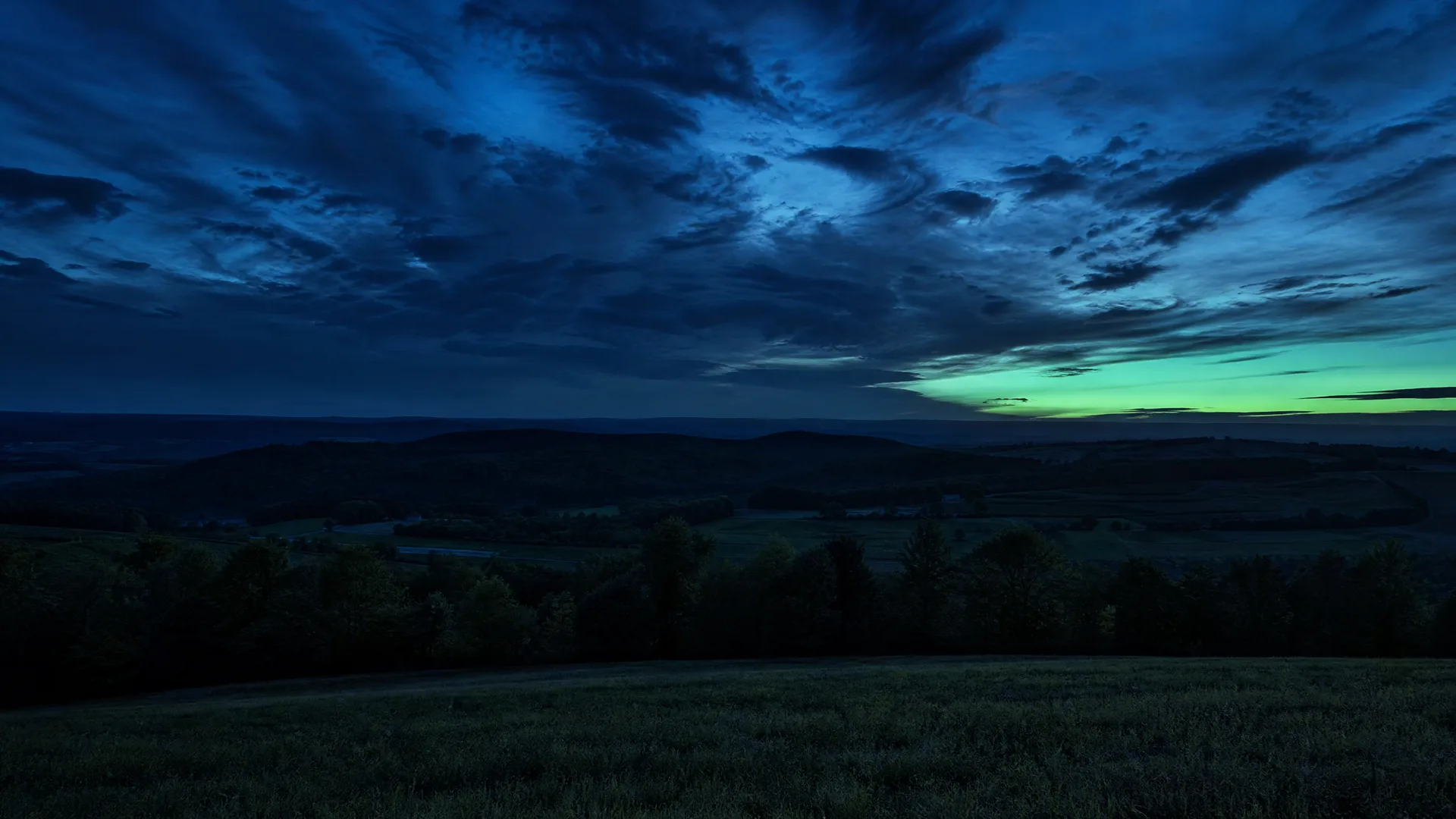 A twilight landscape with a dark grassy field in the foreground, a line of silhouetted trees and low rolling hills beyond, under heavy blue clouds with a green-yellow glow along the horizon at right.
