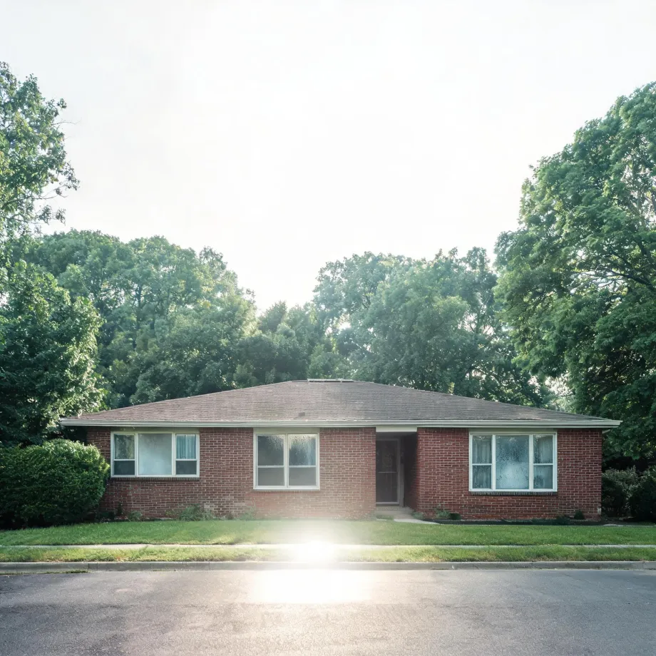 A single-story red brick house with a low hipped shingle roof, white-framed windows and a central door, set behind a mown lawn and street, with large leafy trees behind it under a bright hazy sky.