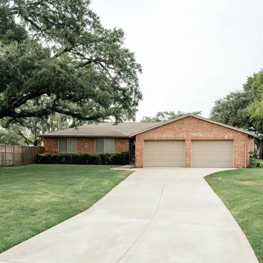 Single-story red brick house with two beige garage doors, a wide concrete driveway, mowed lawn, low shrubs, and a large spreading oak tree overhanging from the left under an overcast sky.