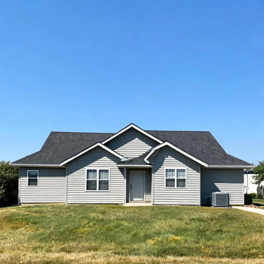 A single-story house with gray horizontal siding, dark shingled gable roof, a light gray front door, and an exterior air conditioning unit, fronted by a green lawn under a clear blue sky.